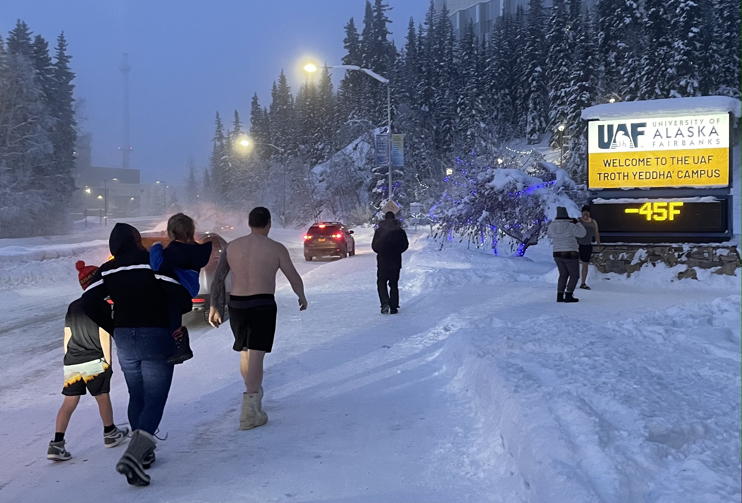 A group of people, some in swim shorts, walk across snow to stand in front of the UAF temperature sign, which reads -45 degrees F.