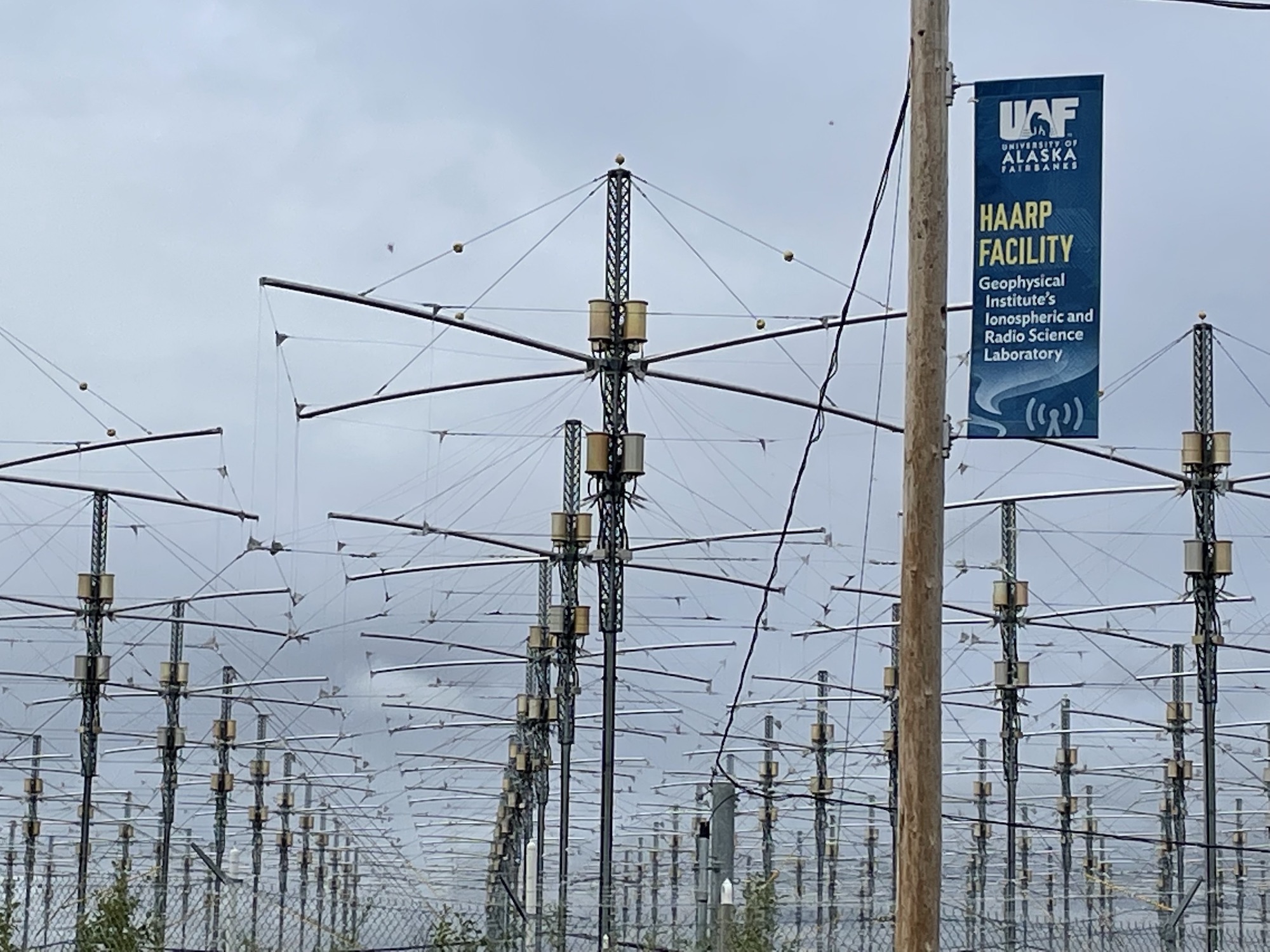 Antenna wires suspended from metal posts are outlined against the sky, while a banner mounted on from a wooden power pole identifies the location as the UAF Geophysical Institute's HAARP facility.