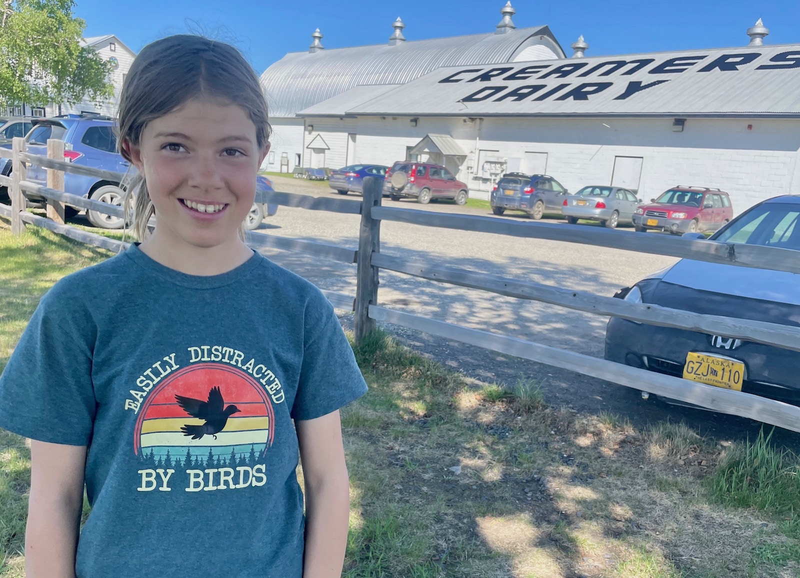 A girl stands near a parking area by restored dairy buildings. Her T-shirt reads "Easily distracted by birds."