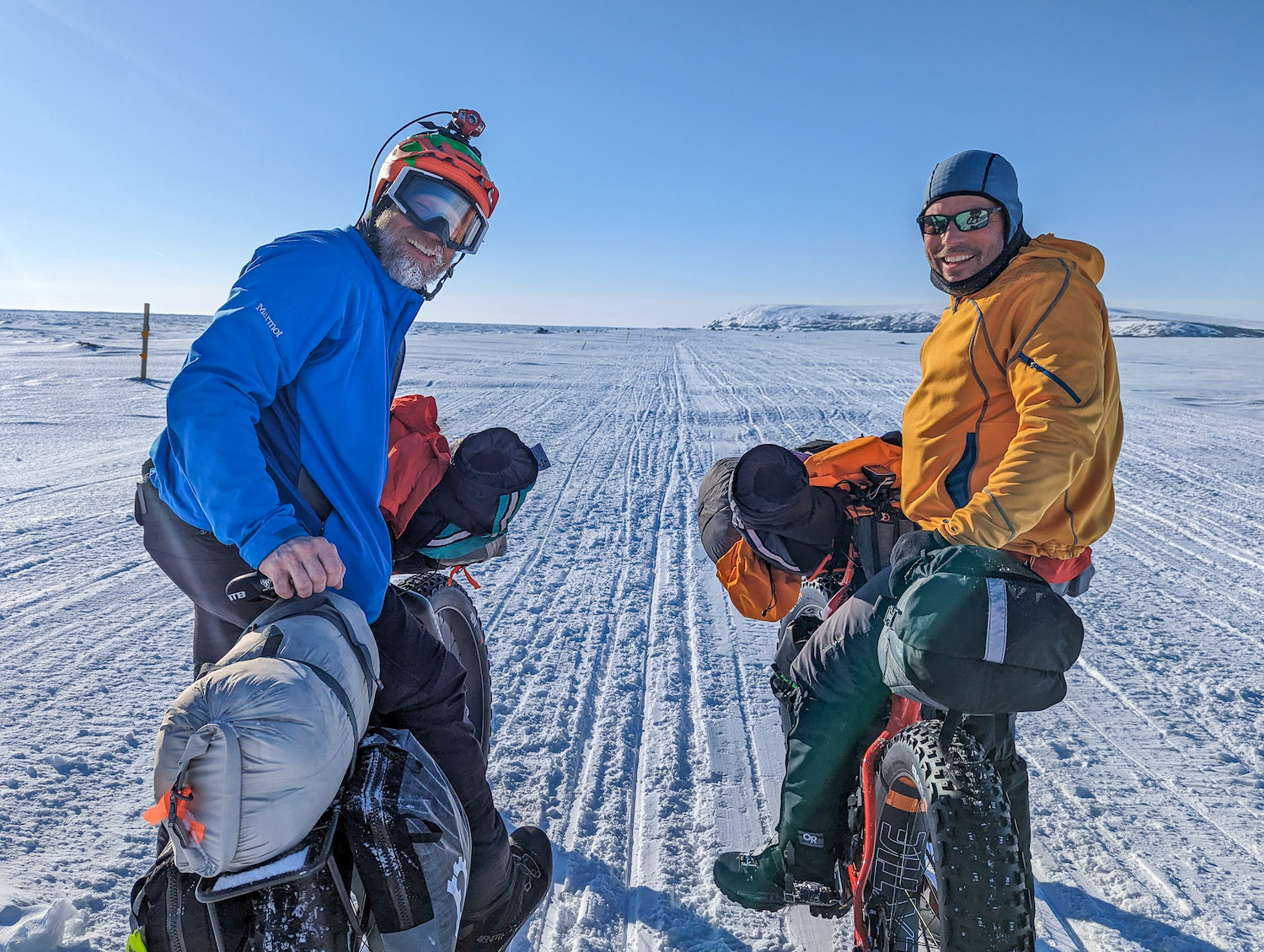 Two men pause astride bicycles on a snow-covered expanse in bright sunlight.