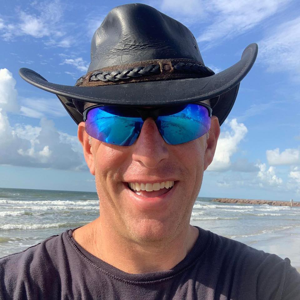 A man in a leather cowboy-style hat and blue reflective sunglasses stands in front of a beach on a sunny day.