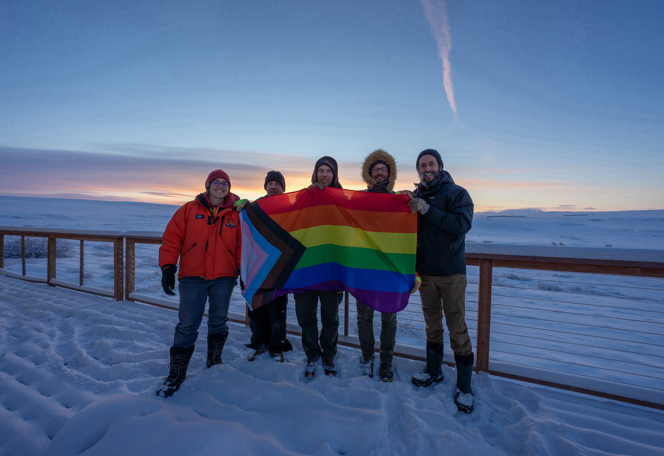 people stand on a deck in the snow holding a pride flag