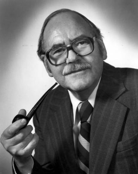 A man with a suit, a tie, glasses and a mustache holds a pipe in a black and white portrait.