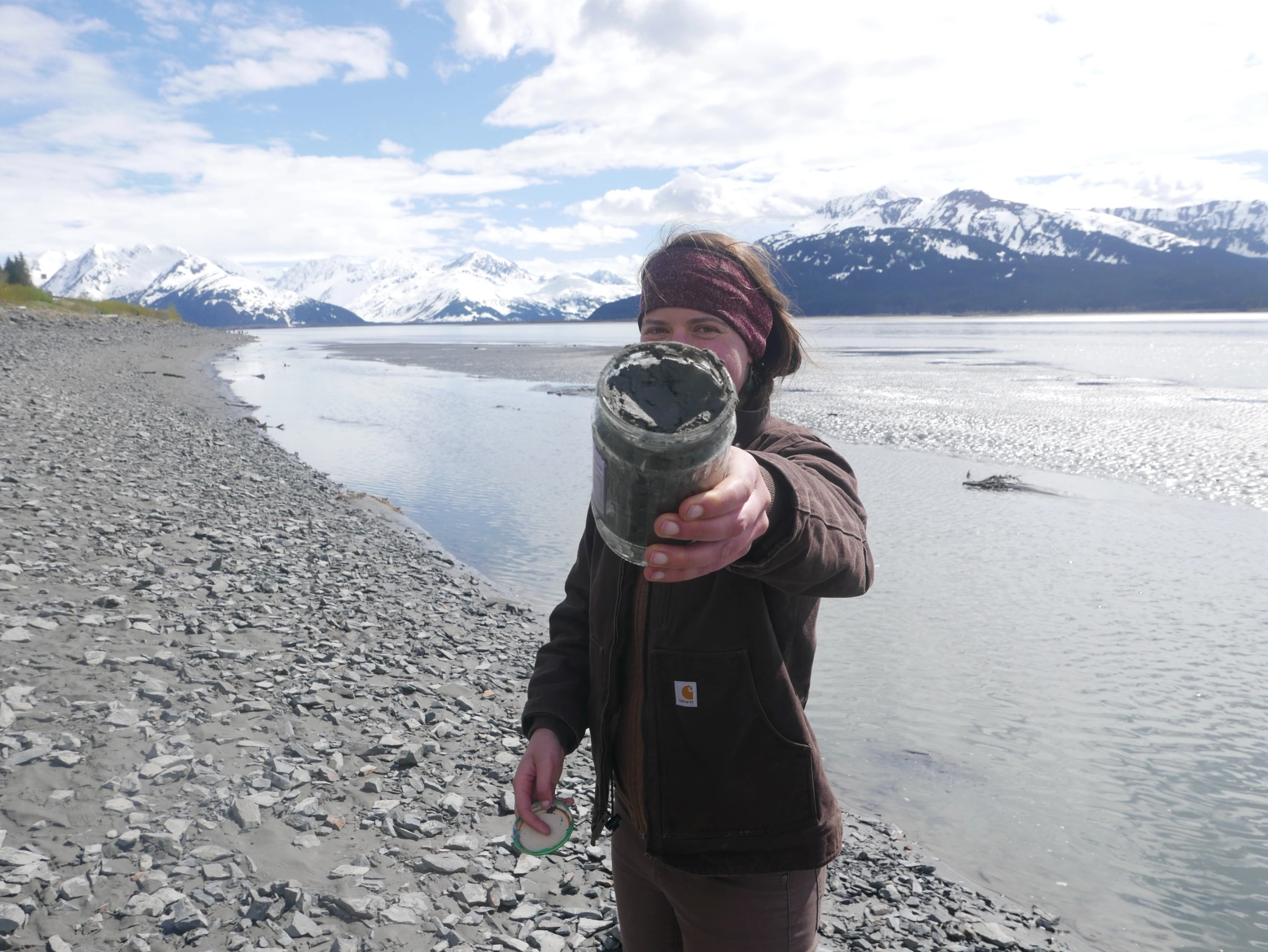 A woman holds up a jar of sediment while standing on a beach with a tidal mudflat, water and snow-covered mountains in the background.