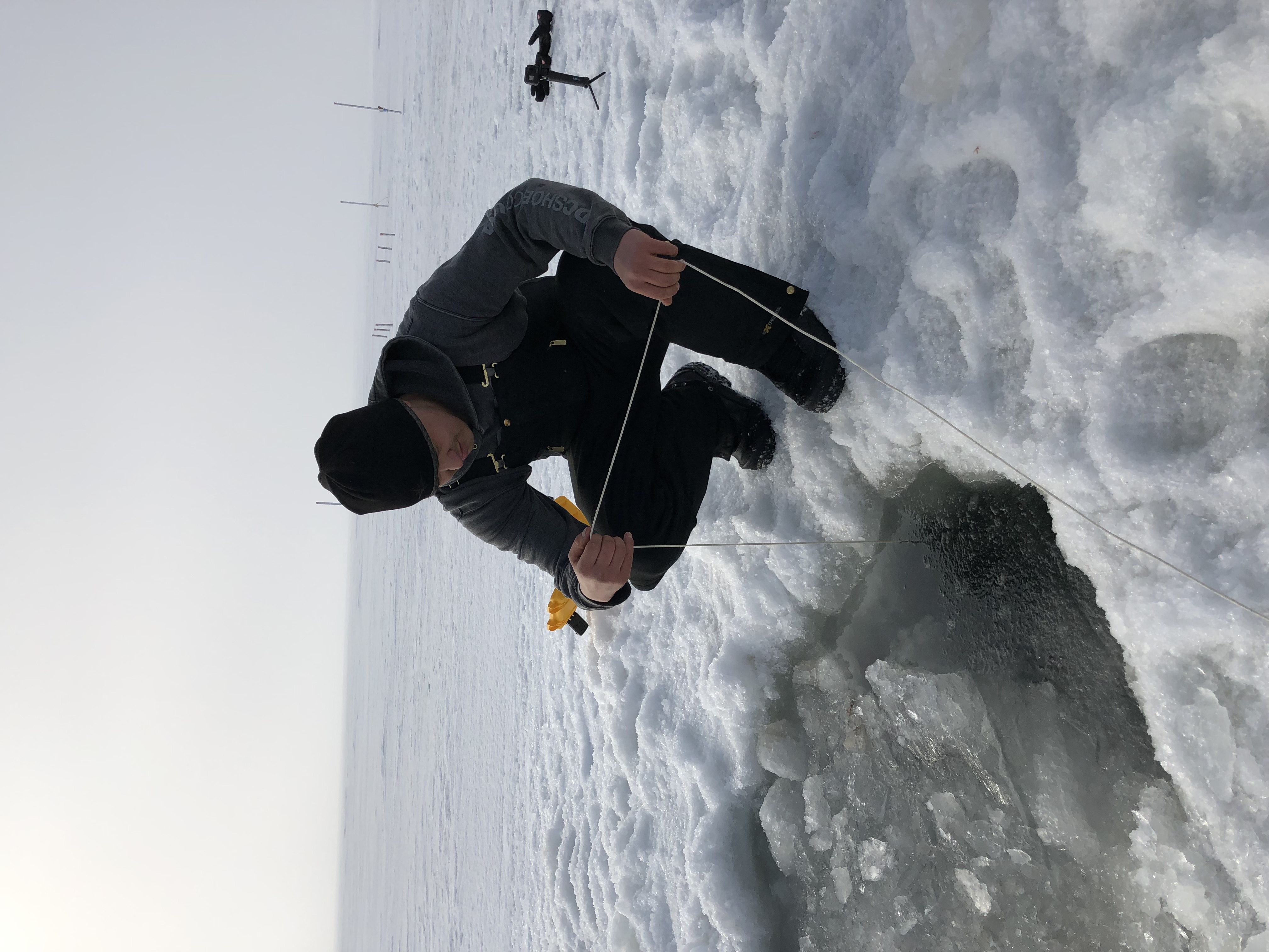 A man holding a line kneels at the edge of a hole in the ice filled with slushy water.