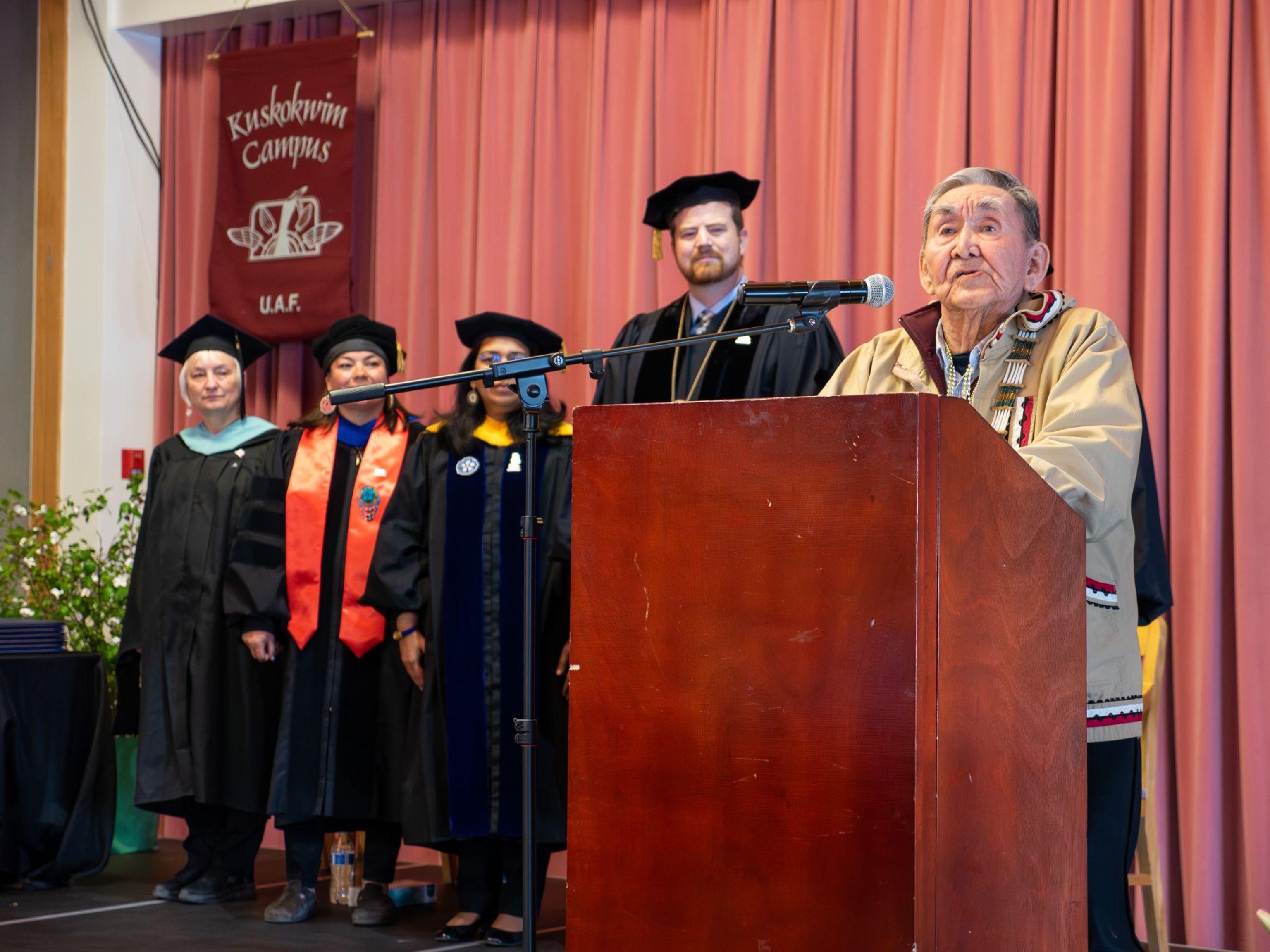 A man wearing a kuspuk speaking from a lectern on a stage. Four people in academic regalia stand behind and to his left, in front of a banner that reads, “Kuskokwim Campus UAF.”