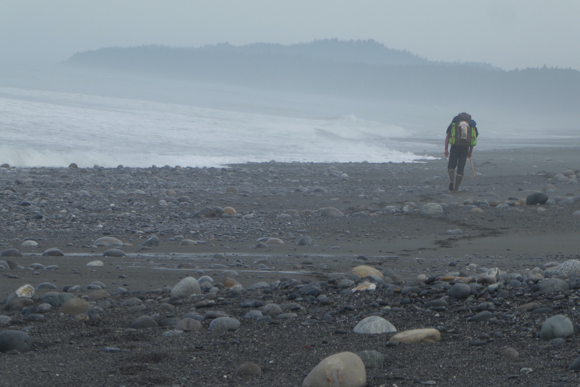 A man carries a backpack along a cobbled ocean beach.