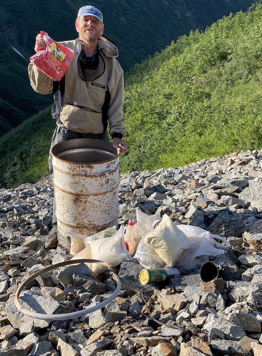 A man holds up a bag of raisins above an opened metal barrel perched on a slope of rocks.