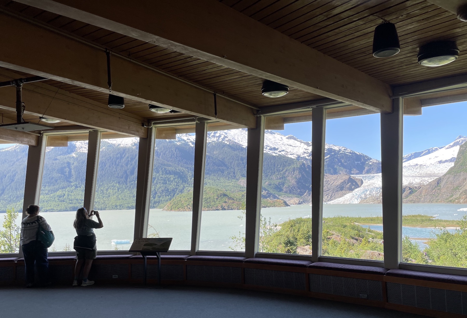 Two women look out an arch of large windows toward a view of a glacier flowing out of snowcapped mountains and into a lake.