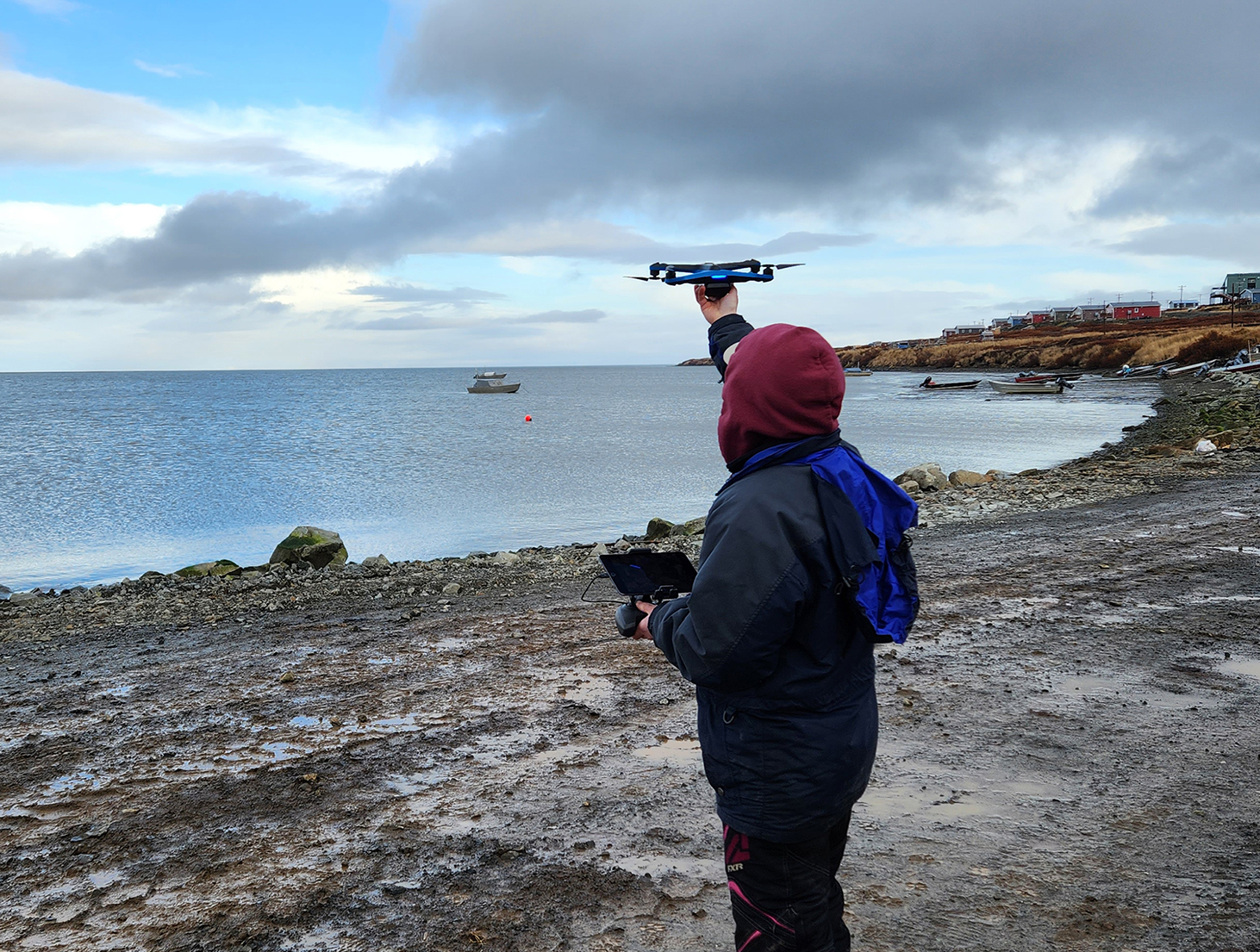 a person in a hoodie holds a drone near the ocean
