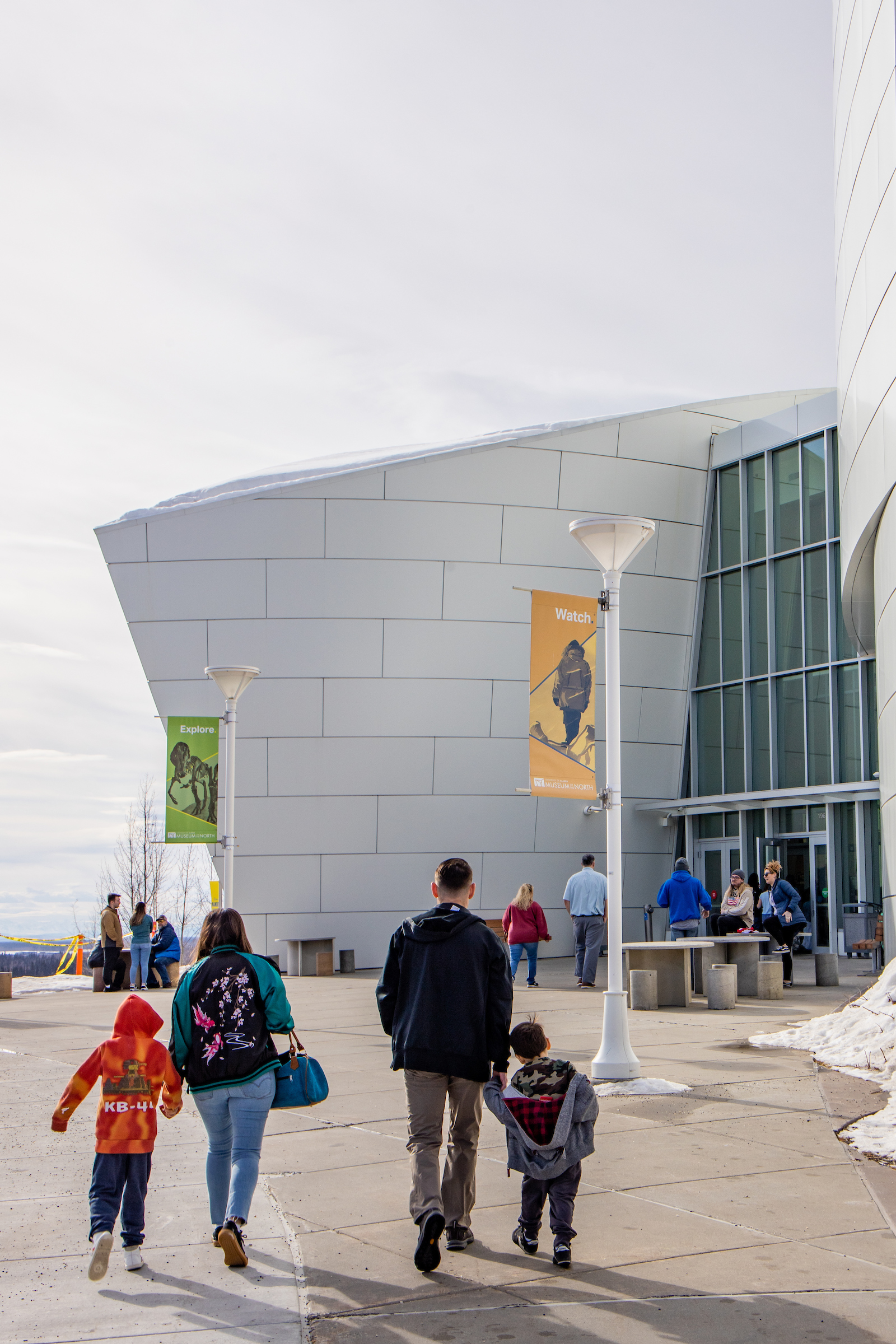 A family of two adults and two children seen from behind approaching the entrance to the UA Museum of the North.