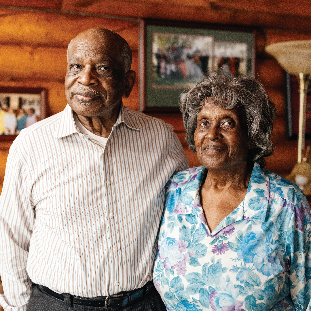 An elderly Black couple stand together in a log cabin interior