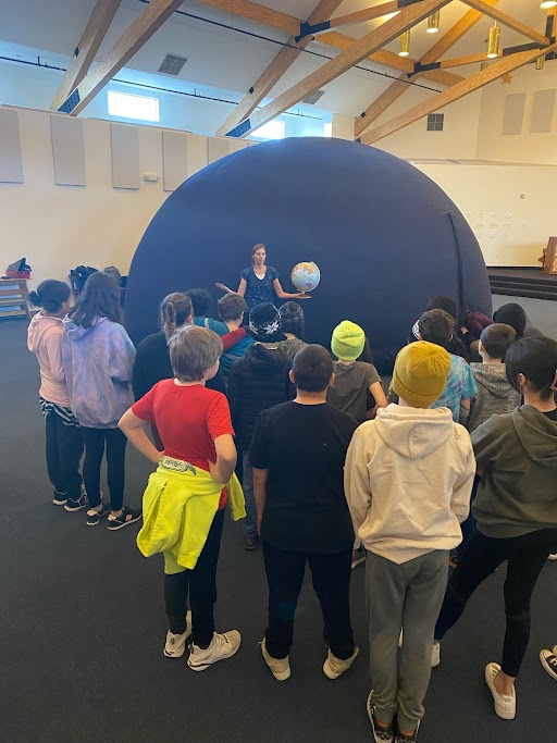 Children gather around the outside of the inflated planetarium to listen to education outreach specialist, Kalee Meurlott, give a talk on planets while holding a globe. 