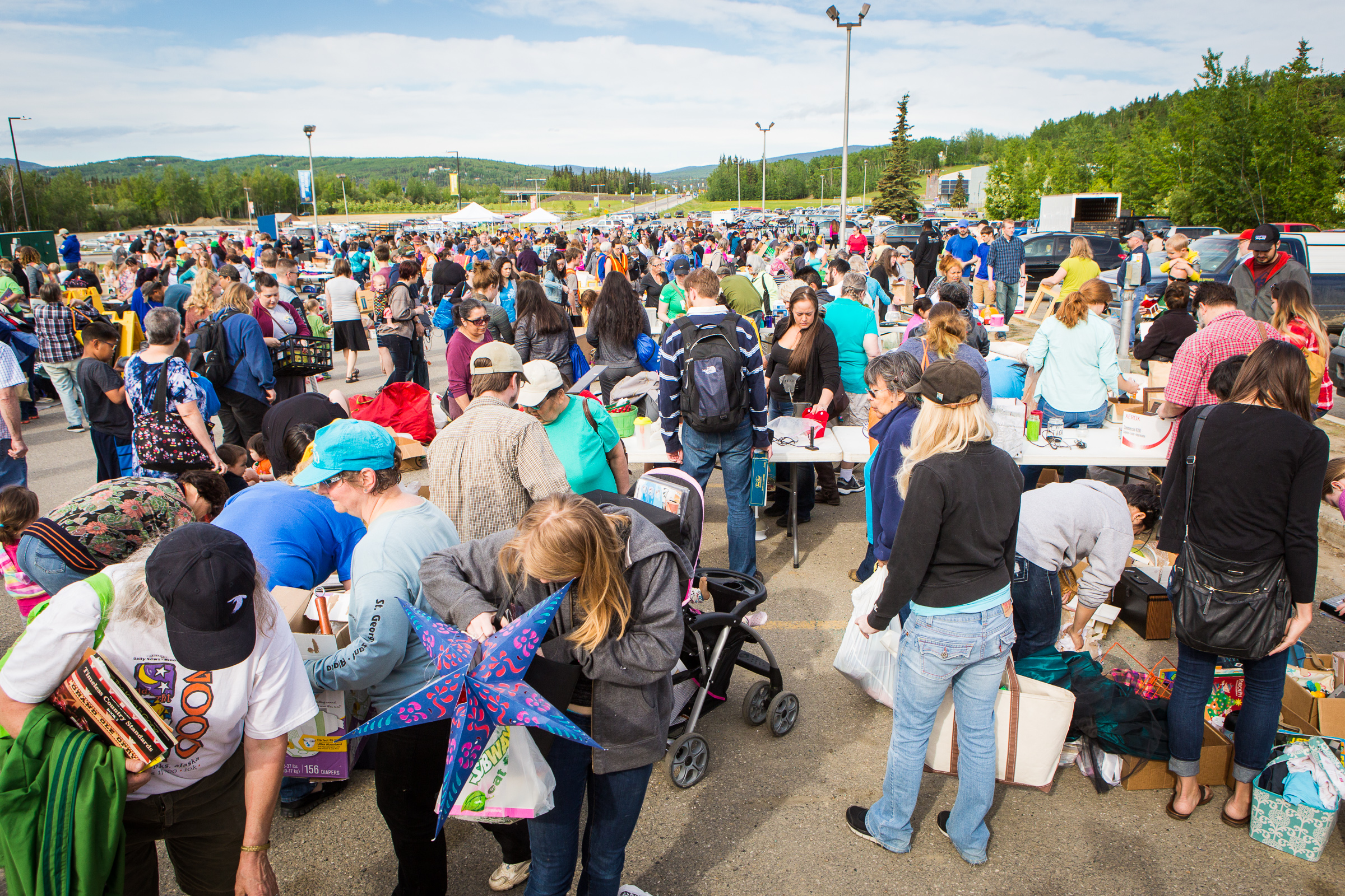 A large crowd of people, many carrying miscellaneous goods, are gathered outdoors in a parking lot with tables and tents.