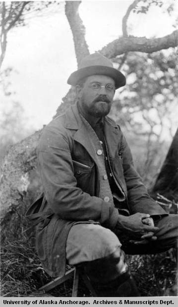 A bearded man with wire-frame glasses, a hat, a heavy canvas coat and boots, sits on a folding stool in a forested area.