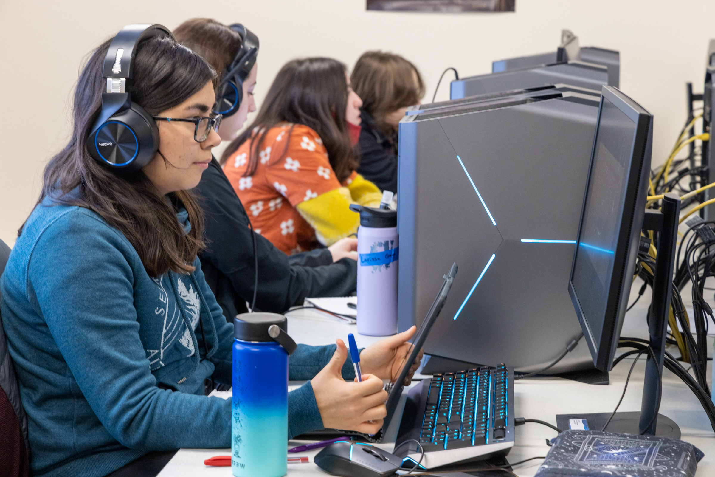 A female college students wearing headphones takes notes while watching a video on at a computer. In the background, three other students work at computers.
