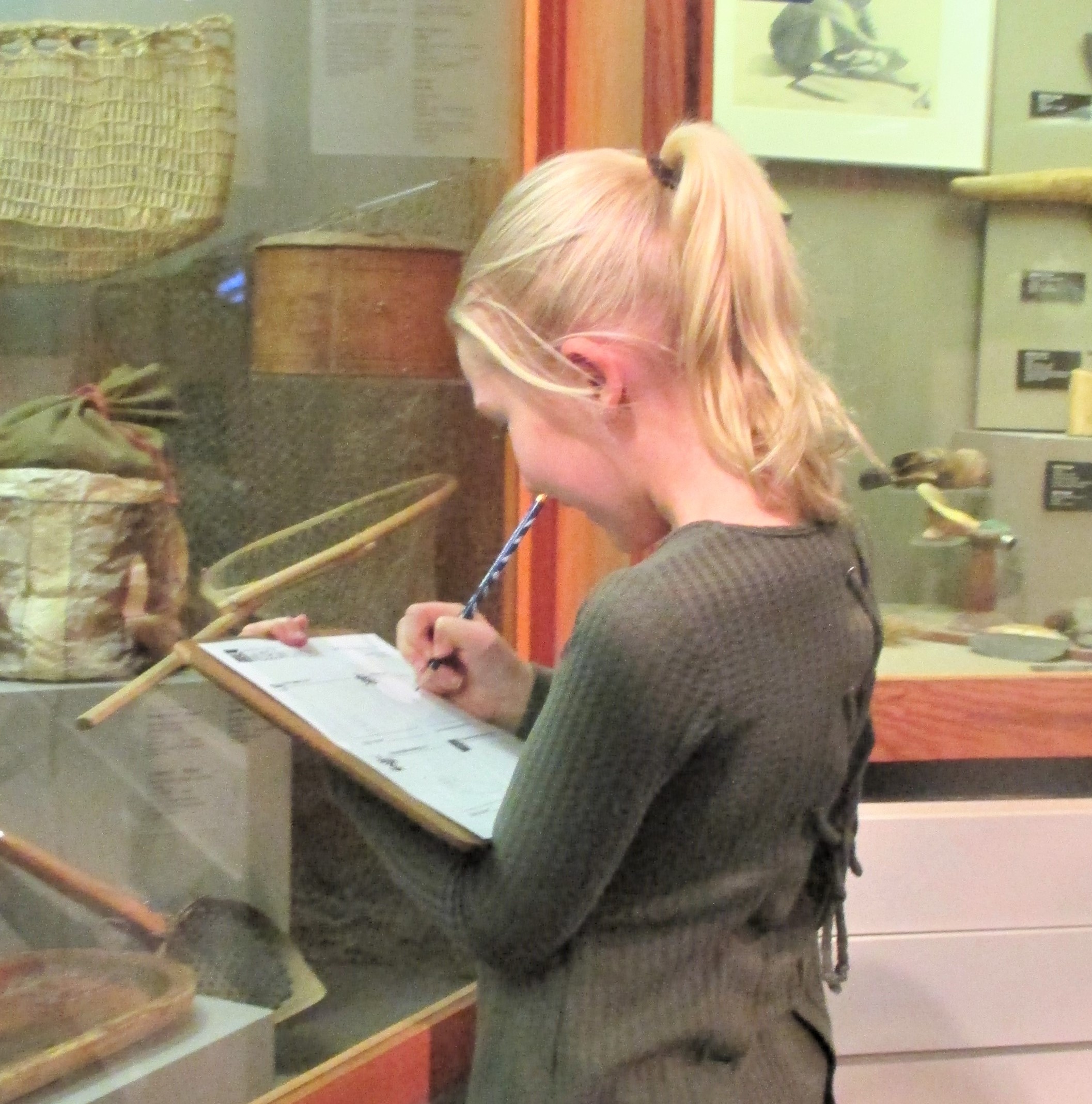 A young child stands in front of a museum display case and uses a pencil to fill out a form on a clipboard.