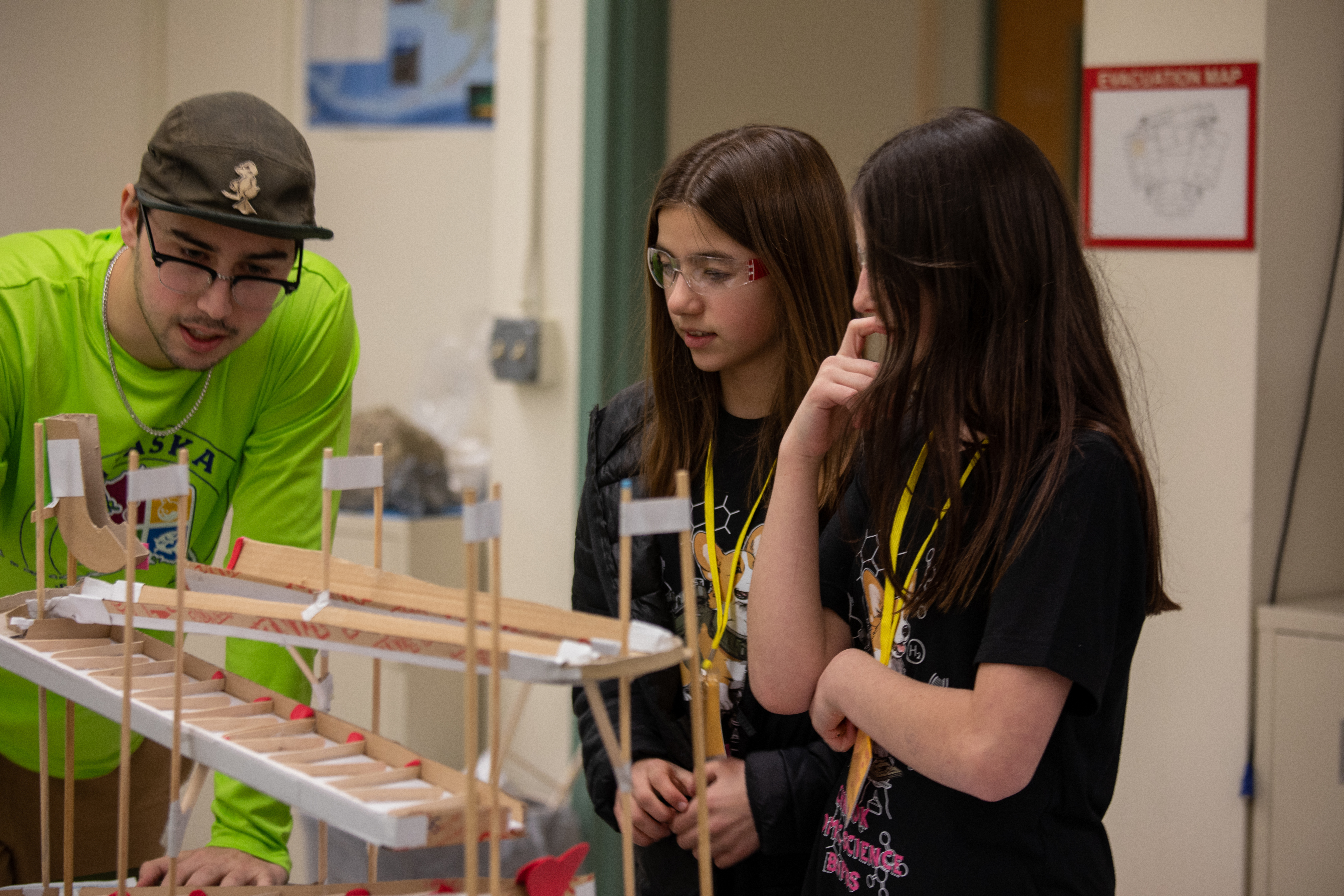 a college student in a green shirt and two middle school students in black team shirts looking at a balsa wood and cardboard model rollercoaster