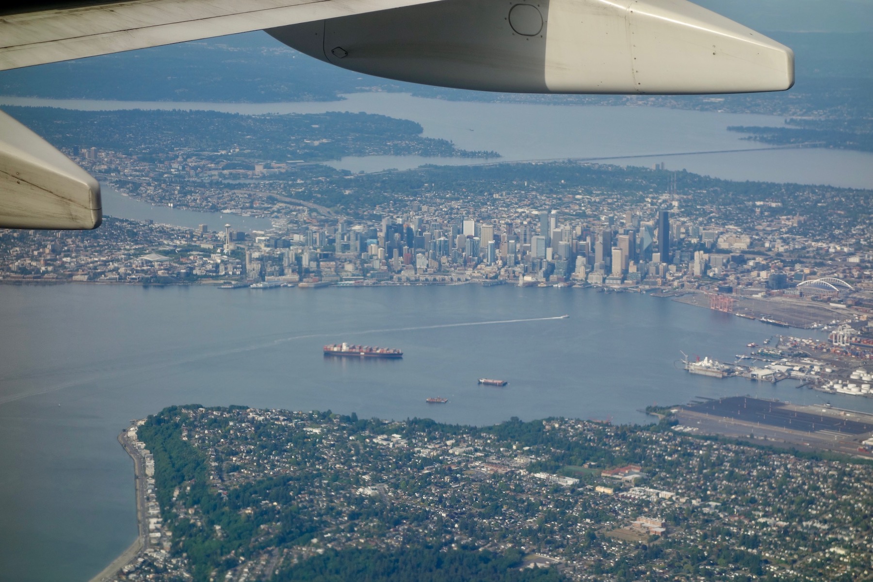 In an aerial photo, Seattle's downtown skyscrapers and its surrounding neighborhoods lie below the flap fairings under a jet wing. Elliott Bay and Duwamish Head are in the foreground, while Lake Washington stretches across the background.