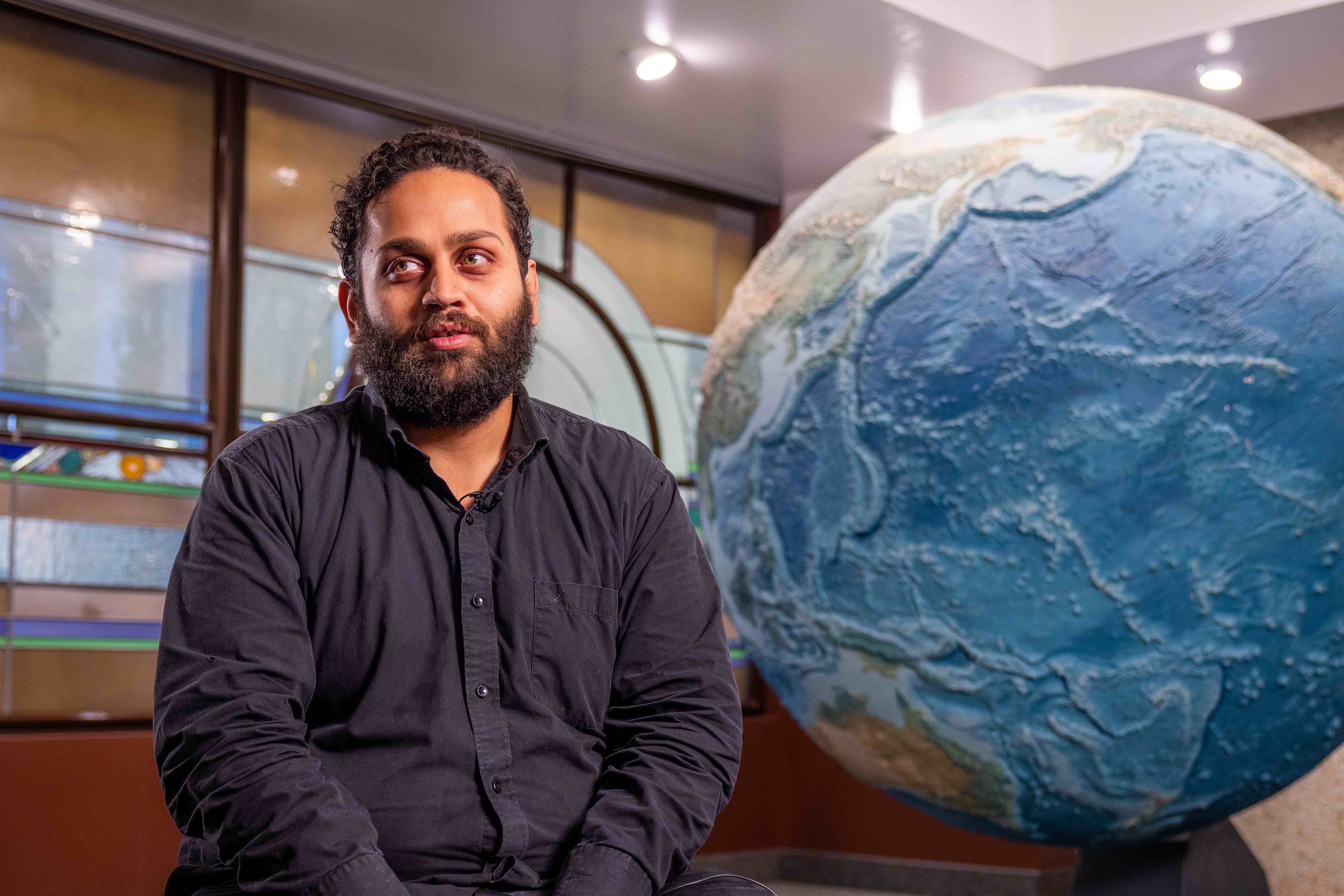 a man with dark hair and beard sits by a giant globe