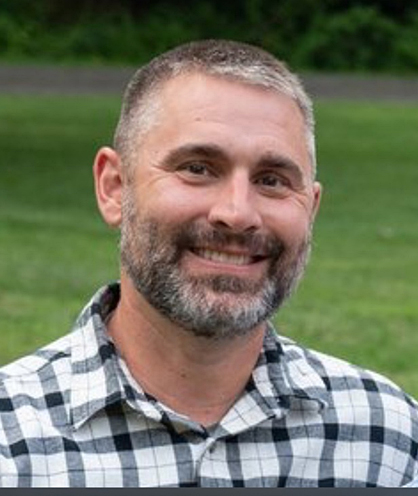 A man with salt-and-pepper-colored hair and a black-and-white checked shirt stands outdoors during the summer.