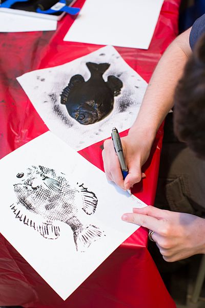 A fish print in black ink on white paper rests on a table beside a dead fish coated in black ink. From out of the frame, a person's hands are seen signing the print with an ink pen.