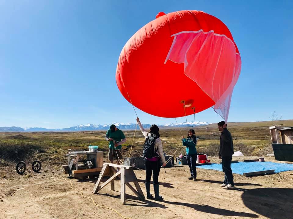 four people holding a red weather balloon with tundra and mountains in the background