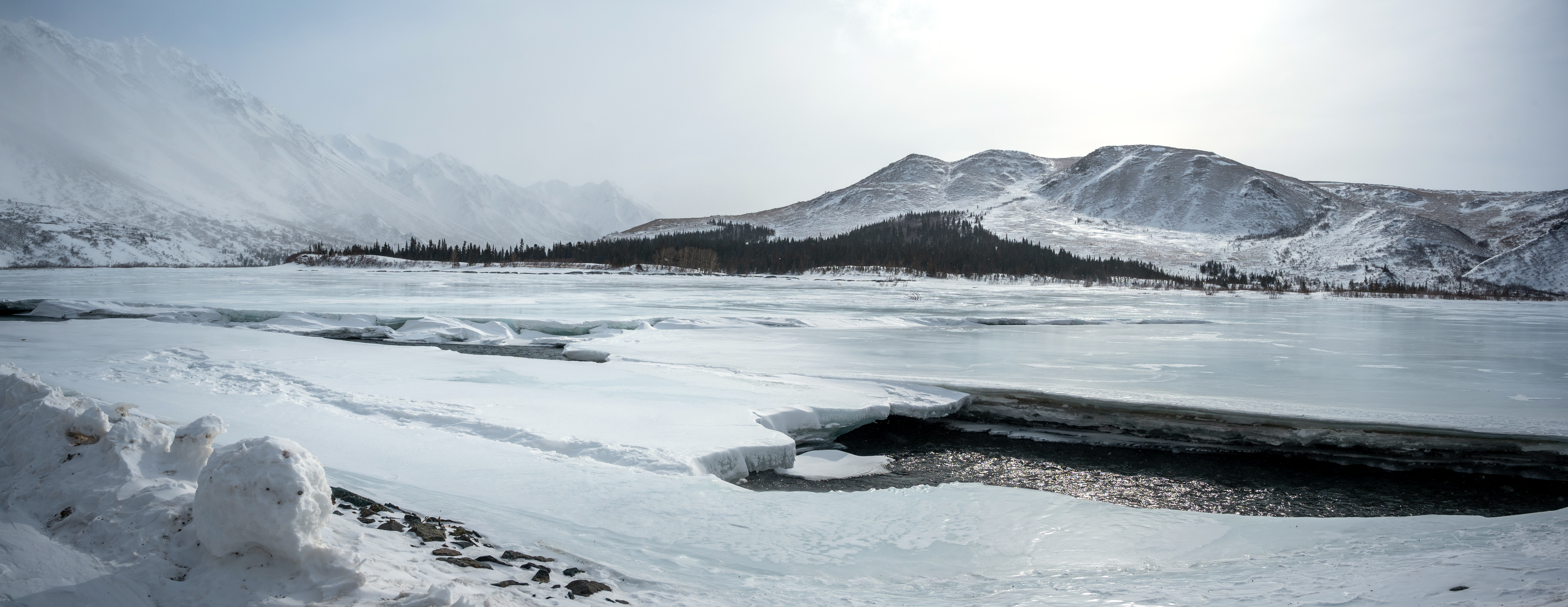 a frozen river with a hole in the ice, with snowy mountains in the background
