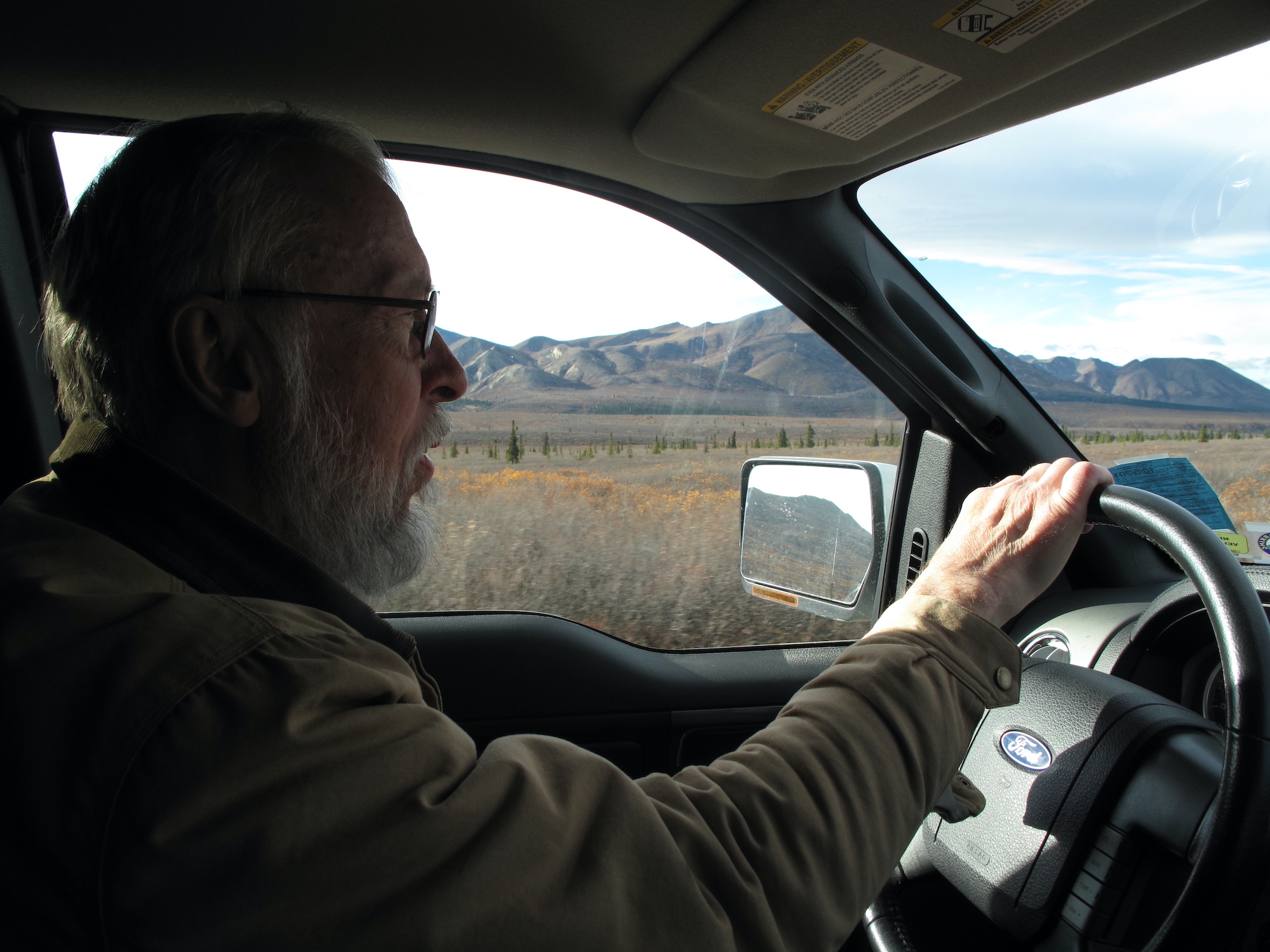 A man with glasses, gray hair and beard drives a vehicle through a fall landscape of tundra and high brush in mountains.