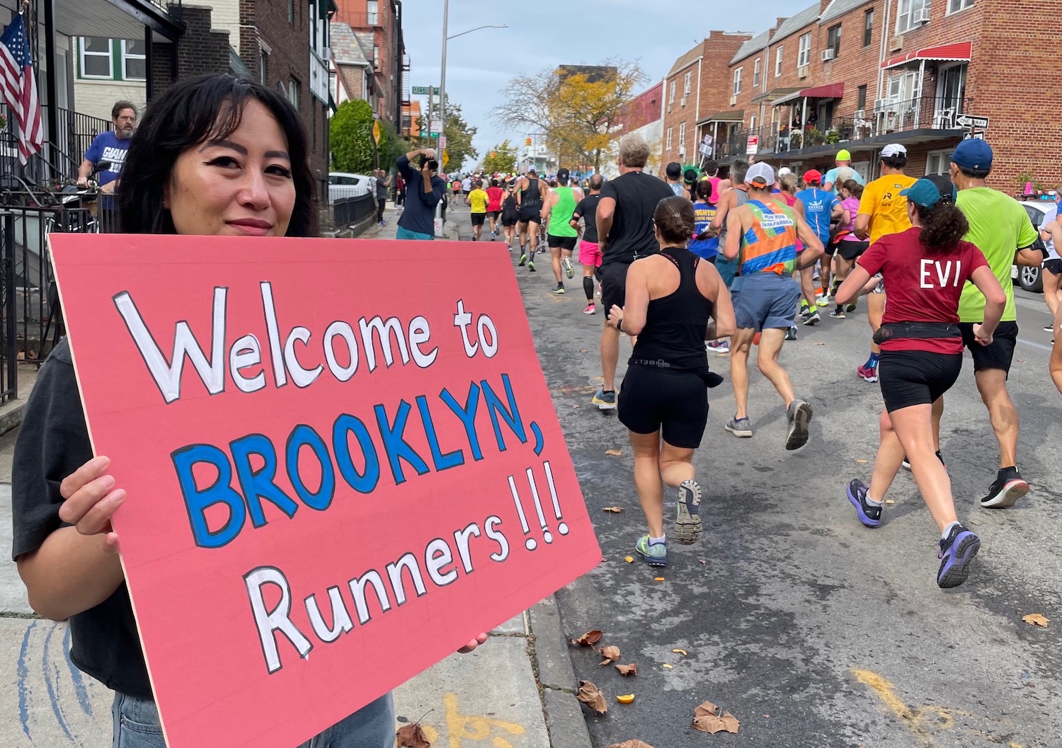 A woman holds a sign saying "Welcome to Brooklyn, runners" as marathon runners pass by on a street.