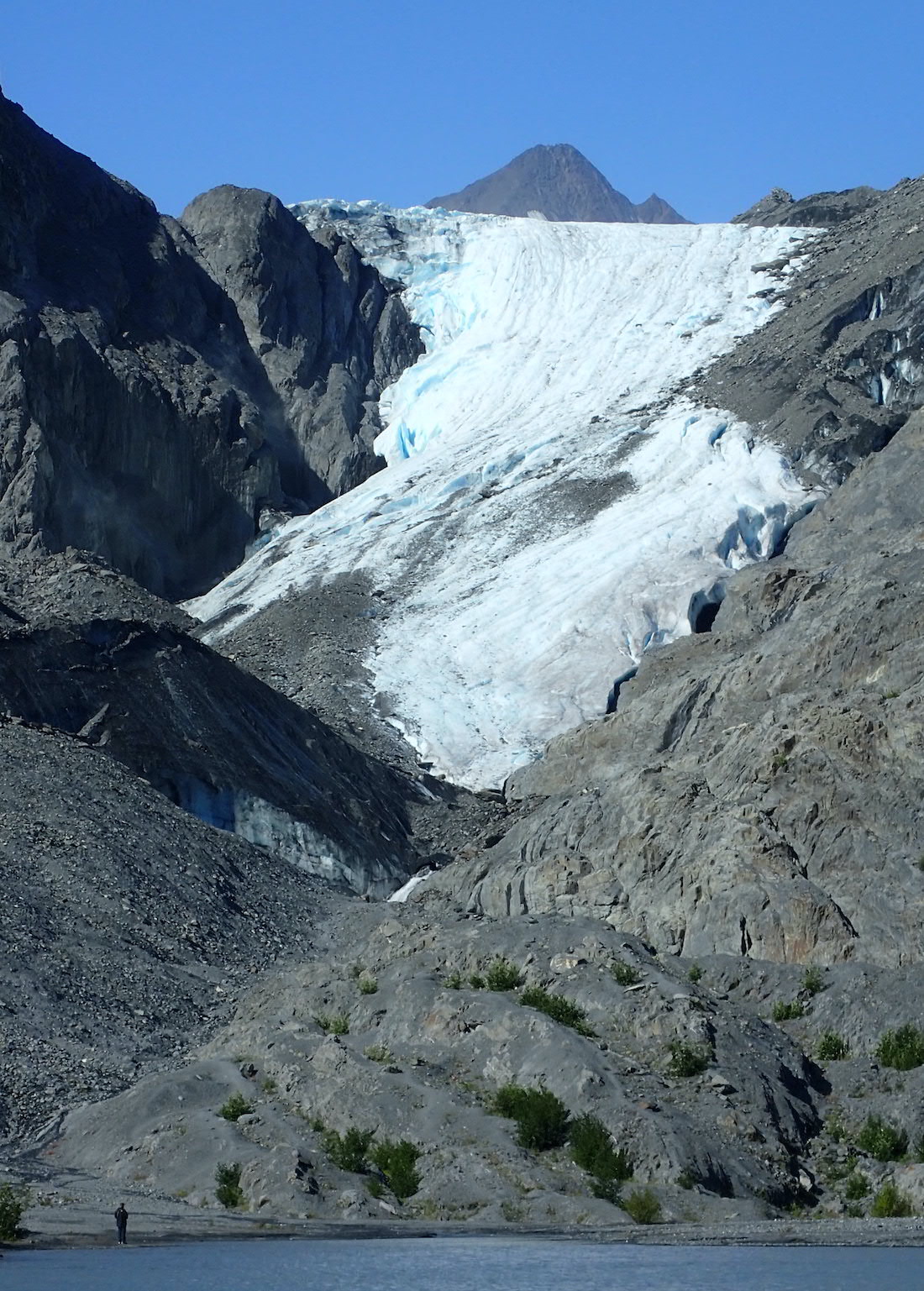A glacier fills a valley between gray rocky slopes. A person stands in the foreground on the shore of a lake.