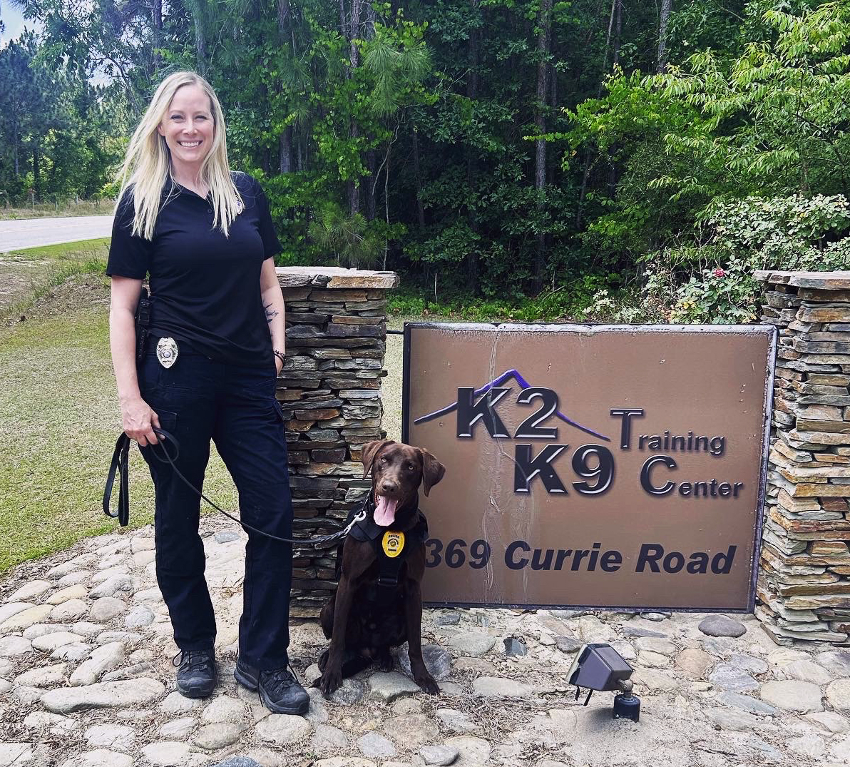 A blonde woman police officer and a chocolate lab police dog stand next to a sign that says "K2 K9 Training Center."