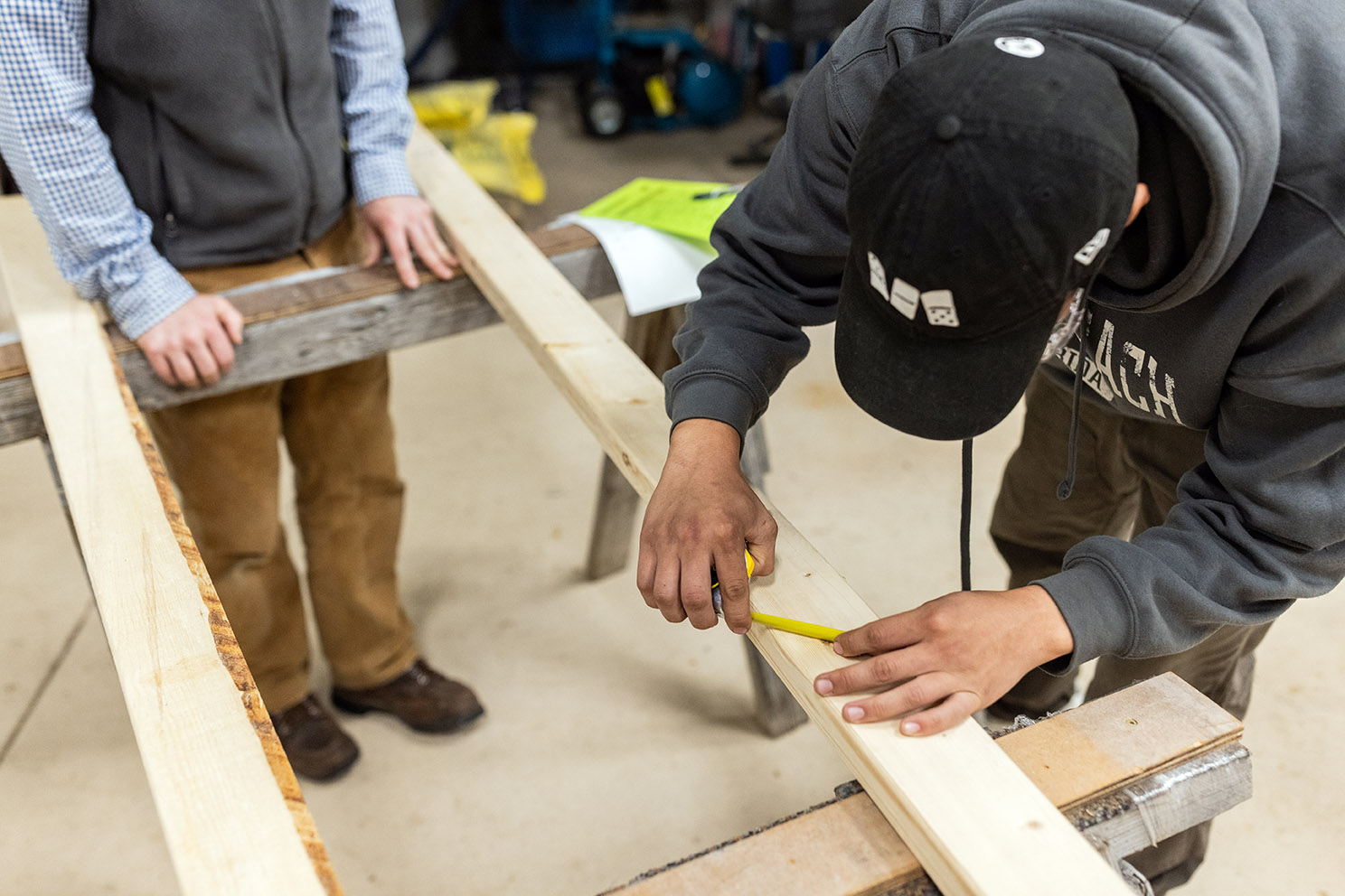 A man leans over a two-by-four piece of lumber with a yellow tape to measure a knot in the wood as another man looks on.