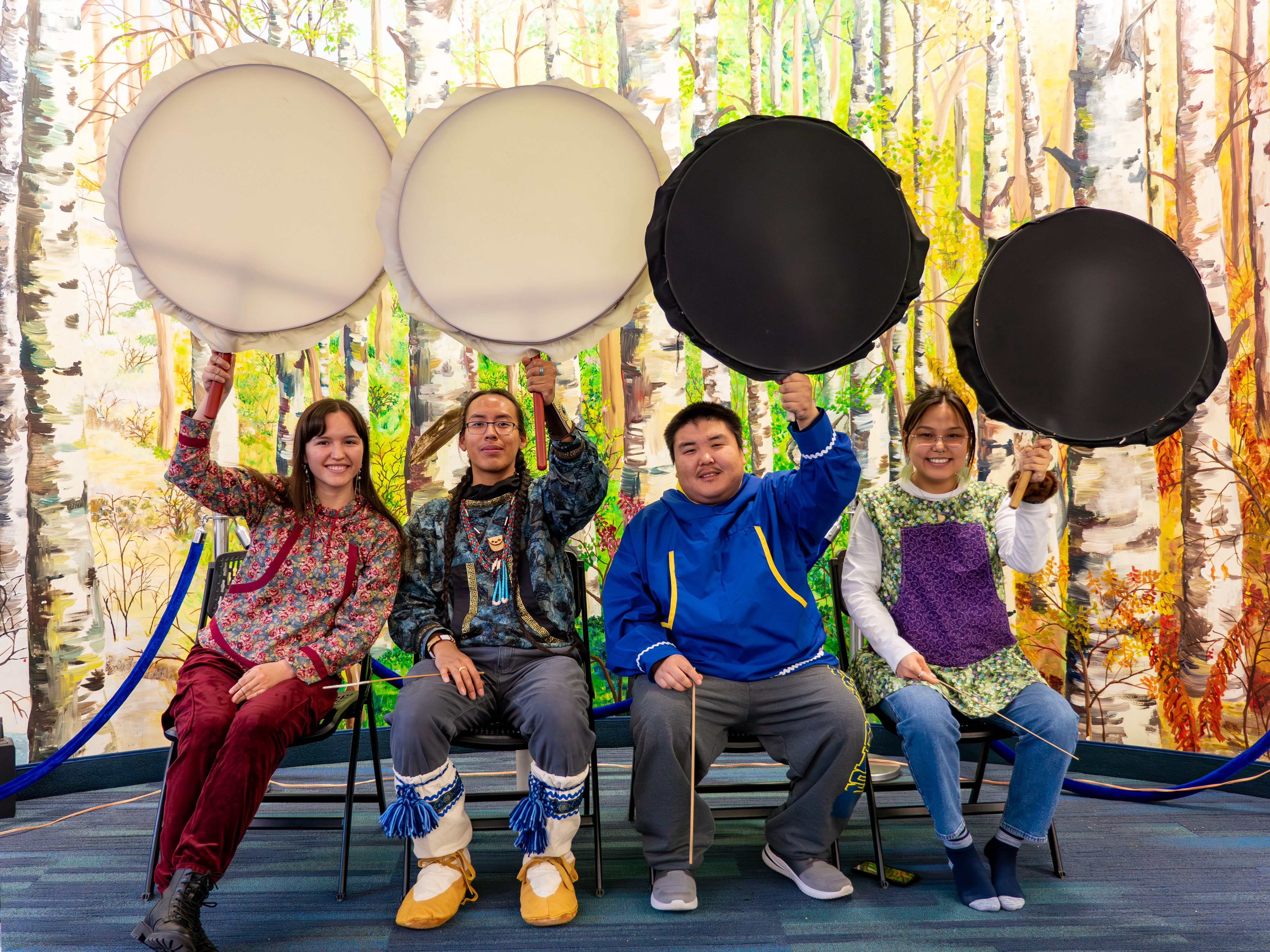 Four people sit on chairs in front of a birch tree mural as they hold up traditional Alaska Native drums.  