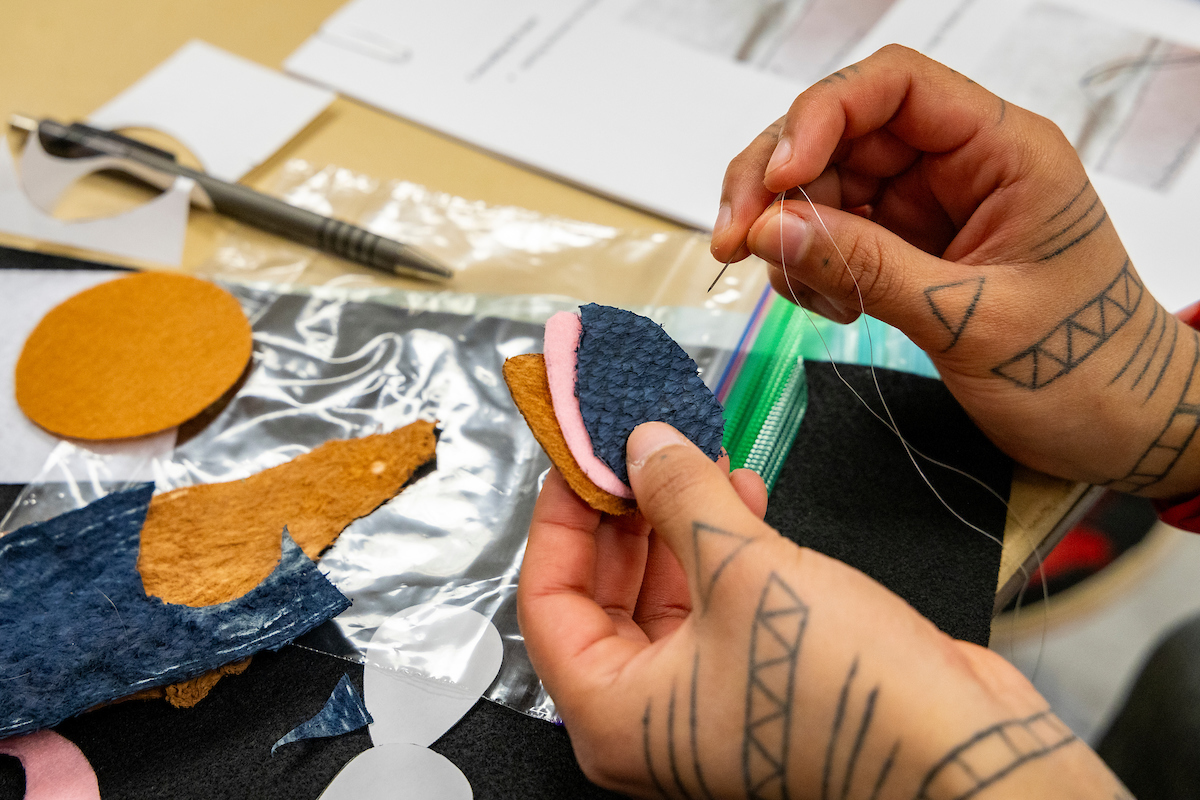 Hangs are seen holding salmon skin and a needle working on a sewing project to create a keychain.