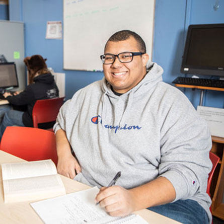 UAF student Kevin Grimes smiles at the camera while seated at a desk with books and course paperwork