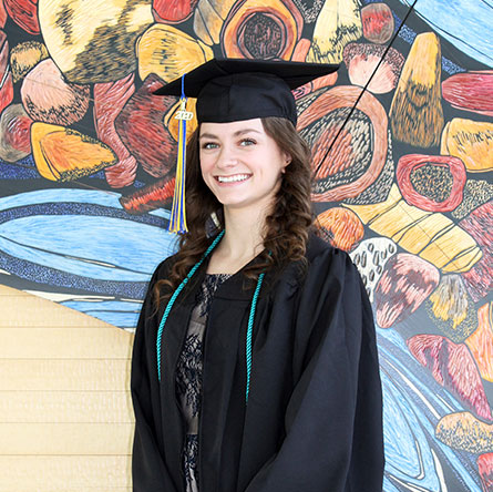 A student in graduation regalia smiles in front of a mural on the UAF campus