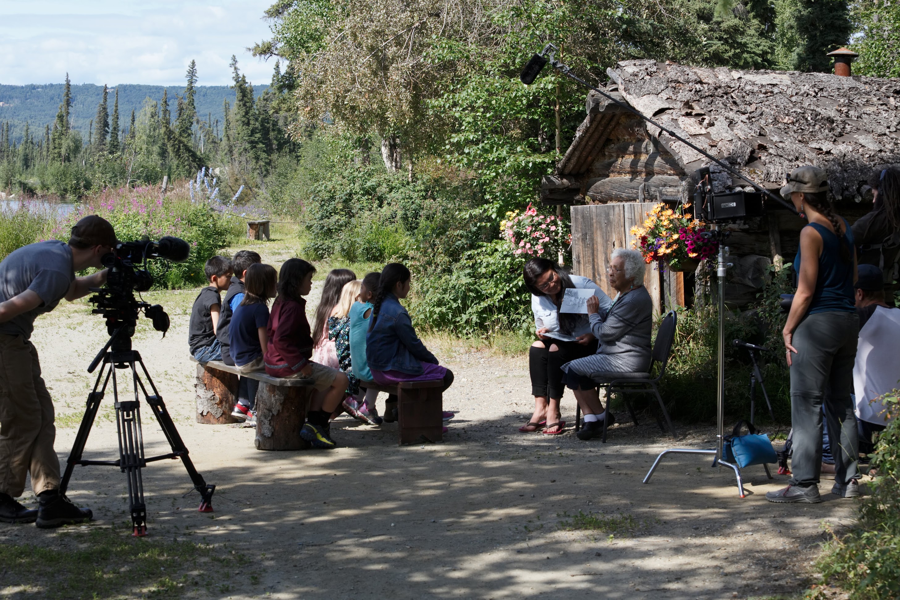 An elder woman tells a story to rows of children outside a small house in the summer. UAF Photo by Maya Salganek