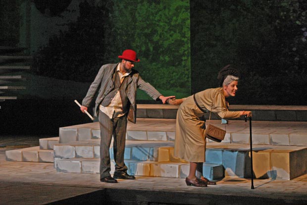 On stage an elderly woman using a cane is walking away from a man holding a cane in his hand. The man reaches for the elderly woman.