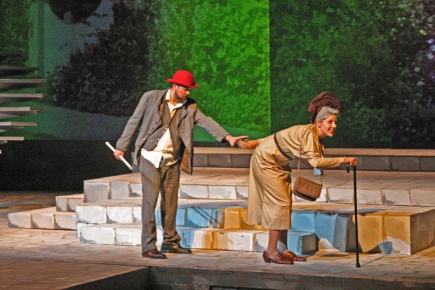 On stage an elderly woman using a cane is walking away from a man holding a cane in his hand. The man reaches for the elderly woman.