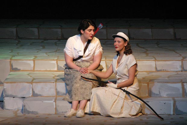 On stage, one woman sits on a stone step with a quiver filled with arrows on her back. Another woman sits to her left, one step below her, holding a bow on her lap.