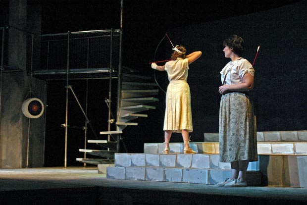 On stage, one woman stands on the second step, pulling back on a bow, preparing to shoot an arrow. Behind her, another woman stands with a quiver filled with arrows on her back, watching intently.