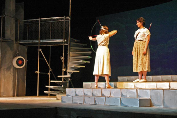 On stage, one woman stands on the second step, pulling back on a bow, preparing to shoot an arrow. Above and behind her, another woman stands with a quiver filled with arrows on her back, watching intently.