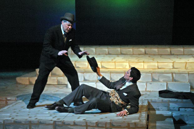 On stage, a man sits on the ground, reaching upward with his right hand, clutching his hat, while another man looms over him. Both appear distraught and angry.
