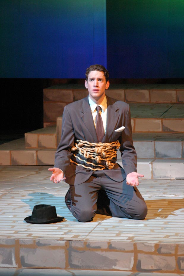 On stage, a man kneels with his waist wrapped in chains and locks, his palms turned upward at waist height.