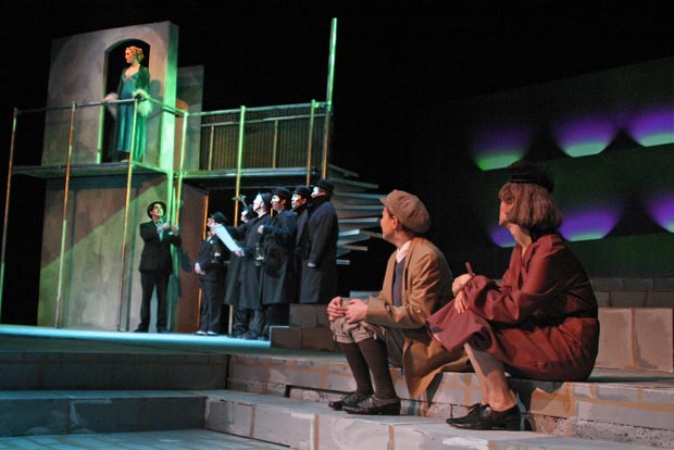 On stage, a man and woman sit on steps, looking to the right at a group of people and women on a balcony.