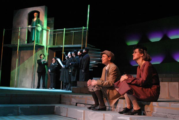 On stage, a man and woman sit on steps, looking to the right at a group of people and women on a balcony.