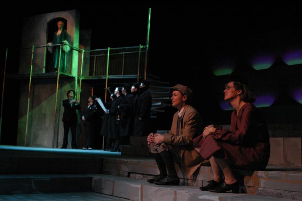 On stage, a man and woman sit on steps, looking out to audience, and a group of people and a women on a balcony is to their right.