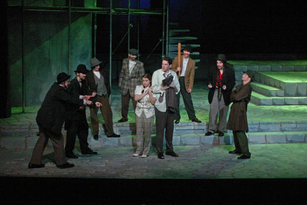 Trapped in the spotlight's glare, a terrified couple confronts a menacing semi-circle of seven men, some brandishing weapons, as uncertainty hangs heavy in the air on stage.