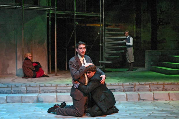 An unconventional embrace: one man kneeling, towering over the other who reciprocates the hug, captured on stage
